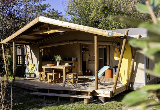 Safari tent Hut Cotton Confort met houten veranda, tuinstoelen, picknicktafel en surfplank in het groen.