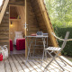 En hyggelig Cabadienne-hytte med terrasse, bord og stole samt to senge indeni, omgivet af grønt.