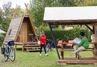 Dos personas junto a una cabaña Cabadienne en Flower Camping La Davière Plage en Francia con bicicletas y árboles.
