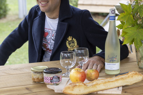 Hombre sentado en mesa exterior con vino francés, copas, baguette, manzanas y rillettes en Cabadienne, Francia.