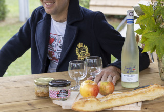 Hombre sentado en mesa exterior con vino francés, copas, baguette, manzanas y rillettes en Cabadienne, Francia.