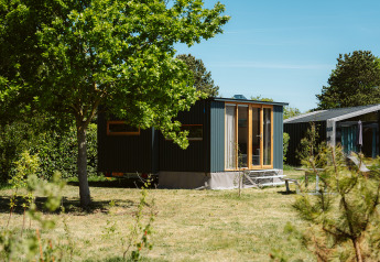 Moderna tiny house Klapkot C20 rodeada de árboles y vegetación bajo un cielo azul despejado.
