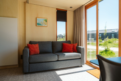 Lichte zithoek in tiny house Klapkot C20 met grijze bank en rode kussens bij De Klepperstee, Nederland.