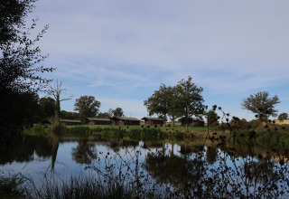View of lake and green fields with lodges at Domaine Bonneblond holiday park in Auvergne-Rhône-Alpes, France.