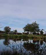 Vista de lago y campos verdes con cabañas en Domaine Bonneblond, parque vacacional en Auvergne-Rhône-Alpes, Francia.