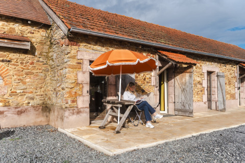 Frau liest unter einem orangefarbenen Sonnenschirm vor einer Steinhütte im Domaine Bonneblond, Frankreich.