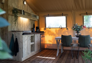Gezellige safaritent met houten keuken, eettafel, groene stoelen en grote ramen met buitenzicht.