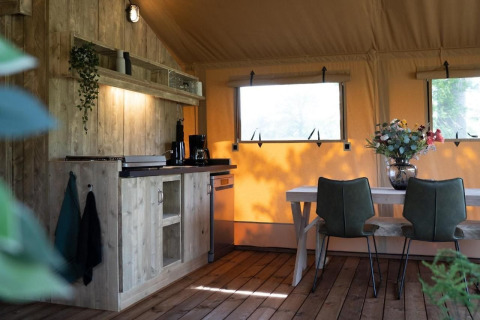 Gezellige safaritent met houten keuken, eettafel, groene stoelen en grote ramen met buitenzicht.
