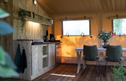 Accogliente interno di tenda safari con cucina in legno, tavolo da pranzo, sedie verdi e finestre luminose.