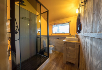 Badezimmer in einem Safarizelt mit moderner Dusche, Toilette und rustikalem Holzdekor bei warmem Licht.