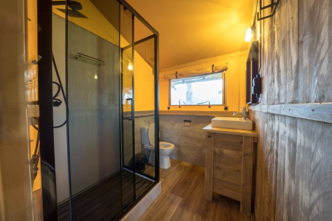Badkamer in een safaritent met glazen douche, houten wastafel en groot raam met buitenzicht en licht.