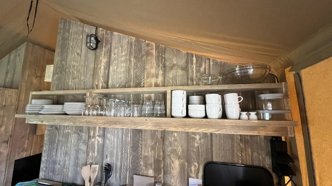 Open wooden shelf in a safari tent kitchen holding neatly arranged plates, mugs, glasses, and bowls.