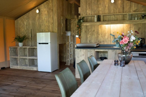 Interieur van een safaritent met houten wanden, eettafel, keukengedeelte en witte koelkast.