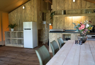 Interieur van een safaritent met houten wanden, eettafel, keukengedeelte en witte koelkast.