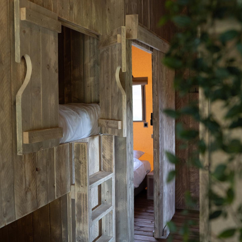 Intérieur de tente safari en bois avec lits superposés, échelle et vue sur une chambre en arrière-plan.