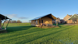 Glampingtent Jannie sur un grand champ vert sous un ciel bleu clair, bâtiments de ferme à l’arrière-plan.