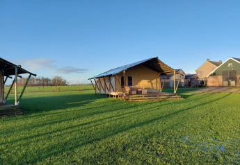 Glampingtent Jannie sur un grand champ vert sous un ciel bleu clair, bâtiments de ferme à l’arrière-plan.