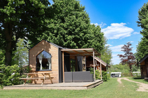 Photo du Tiny House, une petite cabane en bois avec terrasse, banc et verdure, entourée d’arbres.