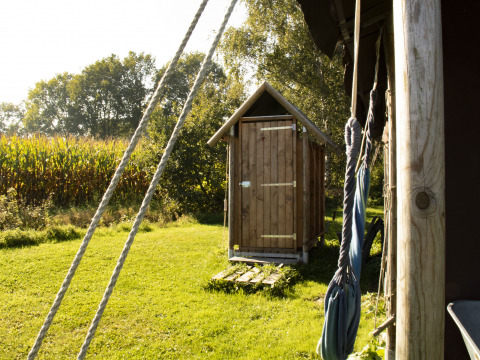 Houten buitentoilet bij Feather Down De Brabantse Hei, omringd door natuur en maïsveld op een zonnige dag.