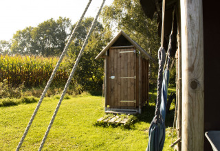 Houten buitentoilet bij Feather Down De Brabantse Hei, omringd door natuur en maïsveld op een zonnige dag.