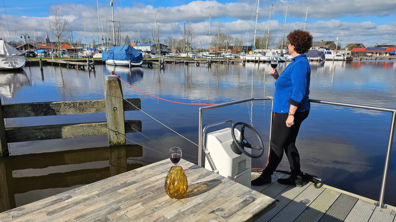 Een vrouw geniet van een glas wijn op het terras bij Marina Heeg, met uitzicht op boten in Friesland.