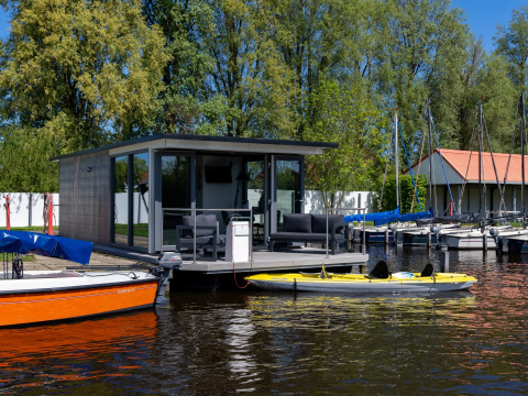 Casa flotante moderna con terraza en Marina Heeg, Friesland, rodeada de botes y árboles bajo cielo azul.