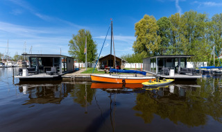 Casa flotante en un puerto deportivo de Frisia, Países Bajos, con barcos y vegetación al fondo.