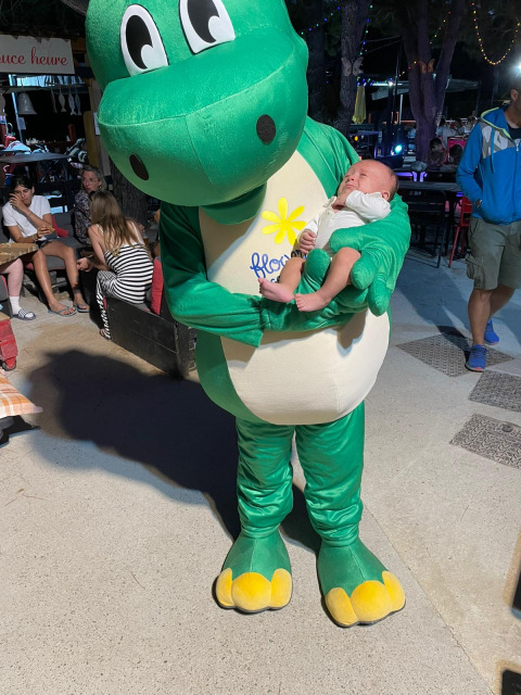 Mascotte in groen dinosauruspak houdt baby vast bij Flower Camping La Rivière, Provence-Alpes-Côte d’Azur.