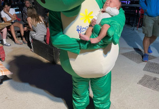 Mascot in a green dinosaur costume holding a baby at Flower Camping La Rivière, Provence-Alpes-Côte d’Azur.