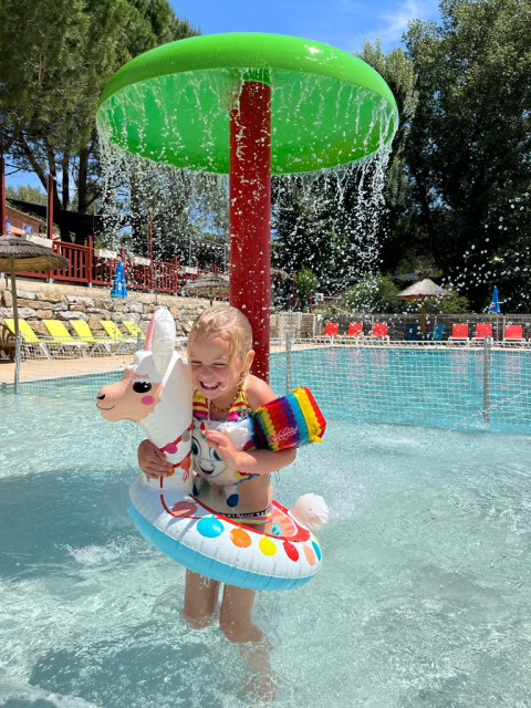 Kind speelt in zwembad met lama-boei onder watersproeier bij Flower Camping La Rivière, Provence, Frankrijk.