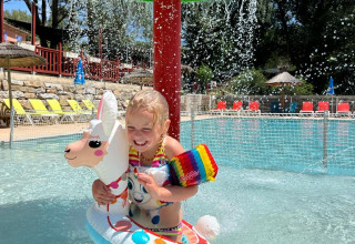 Ein Kind spielt mit einem Lama-Schwimmring unter einem Wasserpilz im Flower Camping La Rivière, Frankreich.
