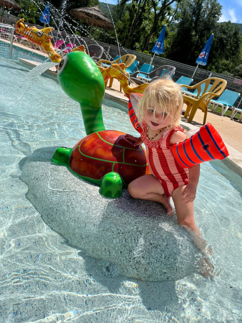 Una niña pequeña juega en un juguete de agua con forma de tortuga en una piscina poco profunda y soleada.