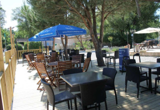 Terraza al aire libre en Flower Camping La Rivière, Provenza, con mesas, sillas y sombrillas bajo los árboles.
