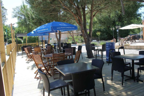 Terraza al aire libre en Flower Camping La Rivière, Provenza, con mesas, sillas y sombrillas bajo los árboles.