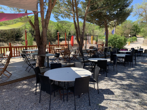 Terraza exterior de Flower Camping La Rivière con mesas y sillas vacías bajo árboles en Provenza, Francia.
