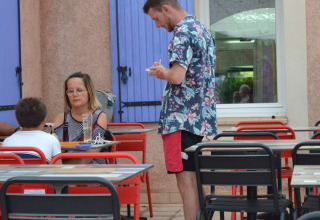 Udendørs café på Flower Camping La Rivière, Provence, med gæster og tjener under hvidt tagnet.