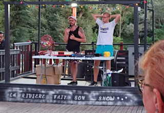 To personer står på en scene ved Flower Camping La Rivière og afholder et udendørs arrangement om aftenen.