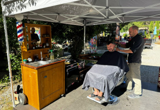 Udendørs barbersalon under telt på Flower Camping La Rivière, Provence, med barber og kunde i stol.