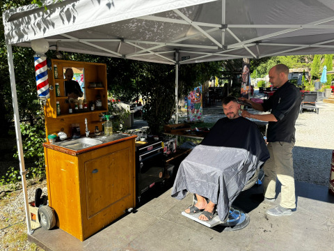 Barbier en plein air à Flower Camping La Rivière, Provence, coiffant un client sous un auvent ombragé.