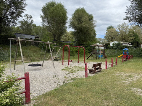 Spielplatz mit Schaukeln, Rutsche und Holzbänken inmitten von Bäumen auf einem Campingplatz in Frankreich.