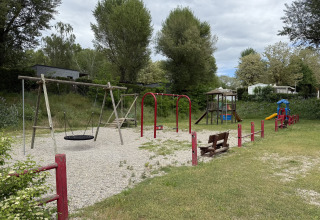 Spielplatz mit Schaukeln, Rutsche und Holzbänken inmitten von Bäumen auf einem Campingplatz in Frankreich.
