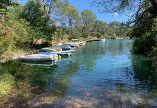 Både fortøjet ved bredden af en fredelig sø omgivet af grønne træer ved Flower Camping La Rivière i Provence.