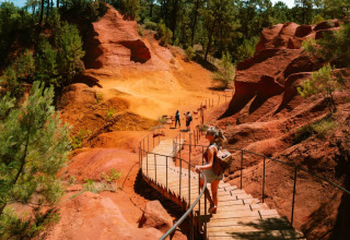 Sentiero di legno tra rocce rosse e pini a Flower Camping La Rivière nella regione Provenza-Alpi-Costa Azzurra.