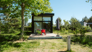 Tiny house with glass front and wooden deck under trees at De Klepperstee Holiday Park in the Netherlands.