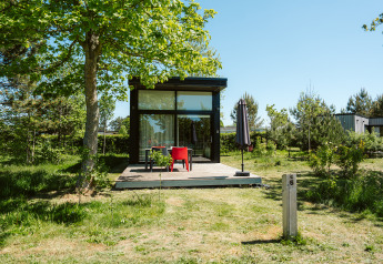 Tiny house with glass front and wooden deck under trees at De Klepperstee Holiday Park in the Netherlands.