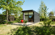 Tiny black wooden house with patio and red chairs in lush greenery at De Klepperstee, Netherlands.