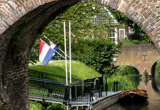 Zicht op een kanaal met aanlegsteiger, Nederlandse vlag en boogbrug in Gelderland bij Feather Down Het Boshuis.
