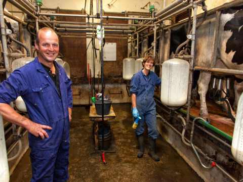 Dos personas con mono azul sonríen en una sala de ordeño con vacas y equipo agrícola en una granja lechera.