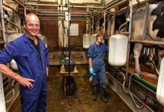Deux personnes en combinaison bleue sourient dans une salle de traite avec des vaches et du matériel laitier.