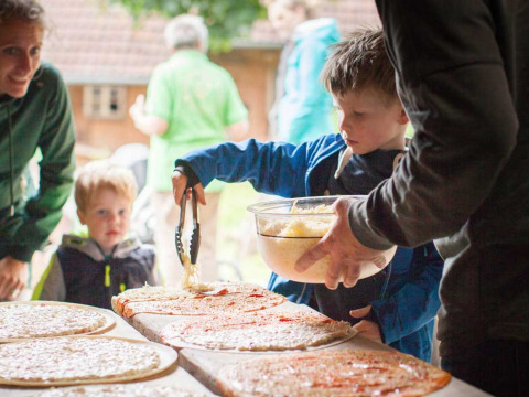 Niños y adultos preparan pizzas caseras al aire libre en Feather Down Hilserhof, un parque de vacaciones en Alemania.